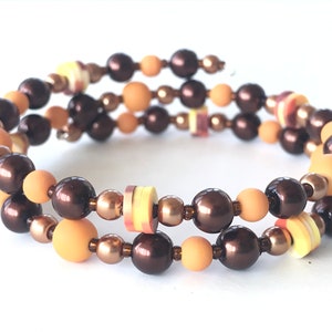 May include: A triple-strand bracelet with brown, peach, and yellow beads. The bracelet is made with a variety of beads, including round, flat, and cylindrical beads.