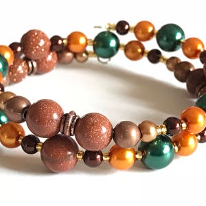 May include: A double-wrap bracelet with brown, orange, and green beads. The bracelet is made with a variety of materials, including glass, metal, and stone.