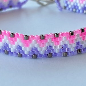 May include: A close-up of a beaded bracelet with a pink, white, and purple color scheme. The bracelet is made with small, round beads and has a silver clasp.
