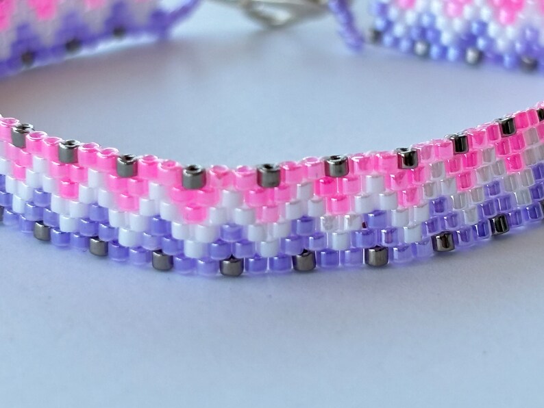 May include: A close-up of a beaded bracelet with a pink, white, and purple color scheme. The bracelet is made with small, round beads and has a silver clasp.