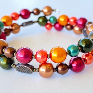 Könnte beinhalten: Ein doppelreihiges Armband mit einer Mischung aus orangefarbenen, roten, grünen und braunen Perlen. Das Armband hat einen Anhänger in Form eines Blattes.