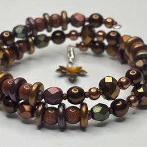 Könnte beinhalten: Ein mehrreihiges Armband mit braunen, goldenen und grünen Perlen. Das Armband hat einen Anhänger in Form eines Blattes.
