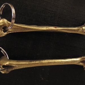 Può includere: Due portachiavi a forma di osso color oro con anelli di salto argentati. I portachiavi sono lunghi circa 7,5 cm.