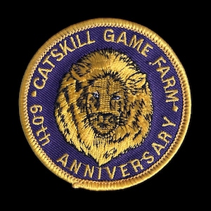 May include: A round patch with a gold embroidered lion's head on a blue background. The text "CATSKILL GAME FARM" and "60TH ANNIVERSARY" are embroidered around the lion. The patch has a gold border.