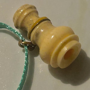 May include: A light beige, decorative bottle stopper with a cylindrical shape and a small, attached gold-colored ring. A teal-colored cord is attached to the ring. The stopper has a series of rounded ridges and a small opening.
