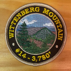 May include: A brown and tan embroidered patch with the text "Wittenberg Mountain #14 - 3,780'" in yellow. The patch features a green mountain with a blue sky and white clouds.