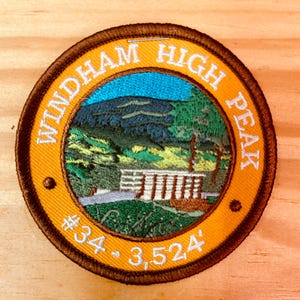 May include: A brown and yellow embroidered patch with the text "Windham High Peak #324 - 3,524'" and a depiction of a mountain range and a building.