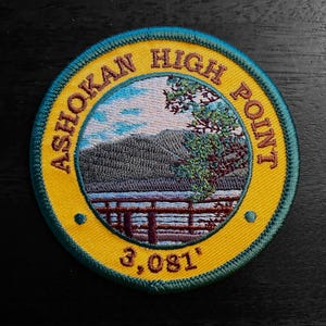 May include: A yellow and green embroidered patch with the text "Ashokan High Point 3,081'" and a depiction of a mountain range, a lake, and a wooden fence.