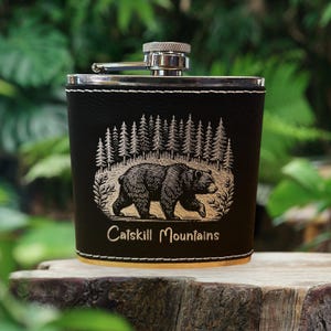 May include: A black leather flask with a silver cap and hinge. The flask features a detailed engraving of a bear walking in front of a forest, with the words "Catskill Mountains" below. The flask is trimmed with white stitching.