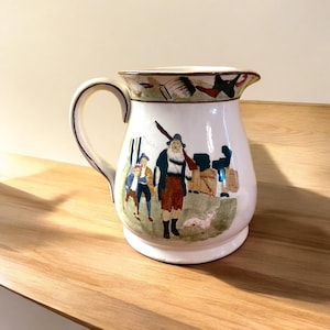 May include: White ceramic pitcher with a brown rim and a painted scene of people and a dog. The scene includes a man with a gun, two children, and a dog.