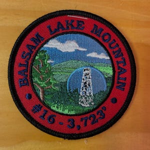 May include: A circular patch with a red border and black trim. The patch features a landscape scene with a mountain, trees, and a tower. The text "BALSAM LAKE MOUNTAIN" arcs around the top. The text "#16 - 3,723" is at the bottom.