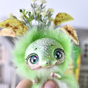 May include: A small, fuzzy green creature figurine with large, expressive teal eyes and a speckled head. The creature features soft, light green fur, small pearlescent limbs, and is adorned with a decorative sprig of white flowers and leaves.