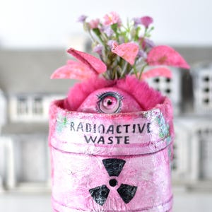 May include: A cylindrical pink container labeled RADIOACTIVE WASTE with a black radiation symbol. The container features a fuzzy pink top with a single realistic eye and a cluster of small pink and purple artificial flowers emerging from it.