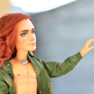 May include: A doll with long red hair wearing a green shirt with gold floral designs, black pants, and a silver chain belt with a seashell pendant.