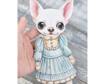 May include: A paper doll of a white chihuahua wearing a blue dress with a yellow collar and lace trim. The doll has a white background and is held by a hand.