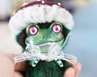 May include: A green frog plush toy wearing a burgundy and white mushroom hat with a white lace trim. The frog has pink eyes and a white lace bow around its neck.
