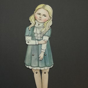 May include: A paper doll of a young girl with blonde hair, wearing a teal dress with a white ruffled collar. The doll has articulated joints and is set against a dark gray background.