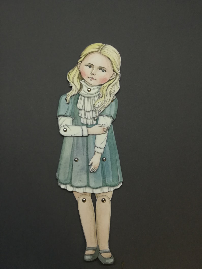 May include: A paper doll of a young girl with blonde hair, wearing a teal dress with a white ruffled collar. The doll has articulated joints and is set against a dark gray background.