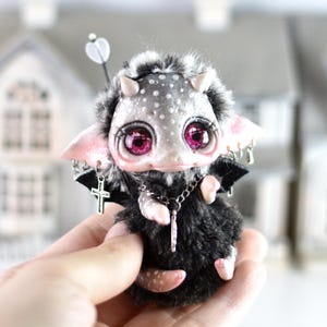 May include: A handmade fantasy doll with a spotted gray face, large pink eyes, and small horns. The doll has a black fur body, pink ears, and silver cross and chain accessories. It is held in a person's hand.