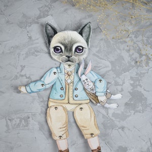 Puede incluir: Una caprichosa mu&ntilde;eca de papel con un personaje de gato con una chaqueta azul claro, pantalones color beige y botas marrones, sosteniendo una mu&ntilde;eca de conejo blanco. El gato tiene una cara gris con ojos morados y est&aacute; articulado con cierres met&aacute;licos.