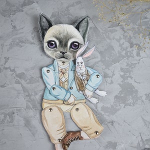 Puede incluir: Una mu&ntilde;eca de papel pintada a mano que representa un gato con una chaqueta azul claro y pantalones color beige, sosteniendo un conejo blanco. El gato tiene grandes ojos morados y lleva una pajarita. La mu&ntilde;eca est&aacute; articulada con broches met&aacute;licos.