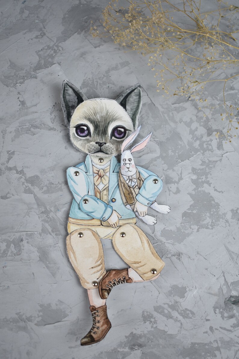 Puede incluir: Una mu&ntilde;eca de papel pintada a mano que representa un gato con una chaqueta azul claro y pantalones color beige, sosteniendo un conejo blanco. El gato tiene grandes ojos morados y lleva una pajarita. La mu&ntilde;eca est&aacute; articulada con broches met&aacute;licos.