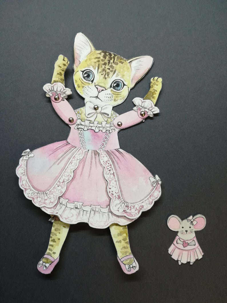 May include: A paper doll of a cat wearing a pink dress with lace trim and a pearl necklace. The cat has brown and black fur and blue eyes. The doll has movable arms and legs. A smaller paper doll of a mouse wearing a pink dress is also included.
