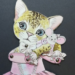 May include: A papercraft cat wearing a pink dress and holding a pink mouse toy. The cat has brown and white fur and blue eyes. The dress has a white lace trim and a bow.