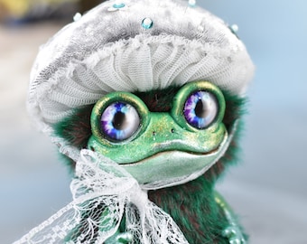 May include: A green frog plush toy with a white lace bow and a silver and white hat with a lace trim. The frog has large blue eyes with purple accents.