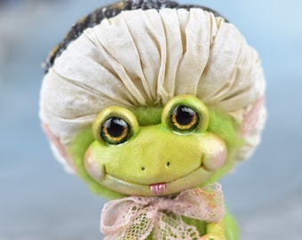 May include: A green frog plush toy with a white fabric shower cap, pink lace ribbon, and large brown eyes. The frog is smiling with its tongue sticking out.