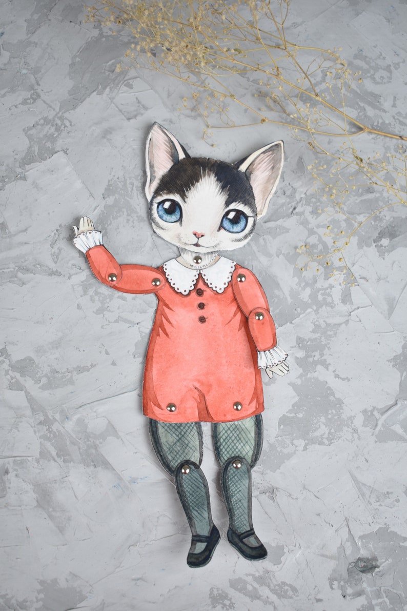 May include: A paper doll of a cat wearing a red dress with a white collar and black tights. The doll has blue eyes and black and white fur. The doll is posed with one arm raised and is on a grey background.