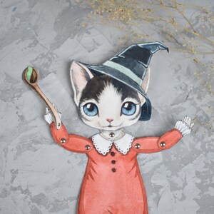 May include: A paper doll of a cat wearing a witch's hat and a red dress, holding a wooden spoon with a green ball on it. The doll is on a grey background with dried flowers.