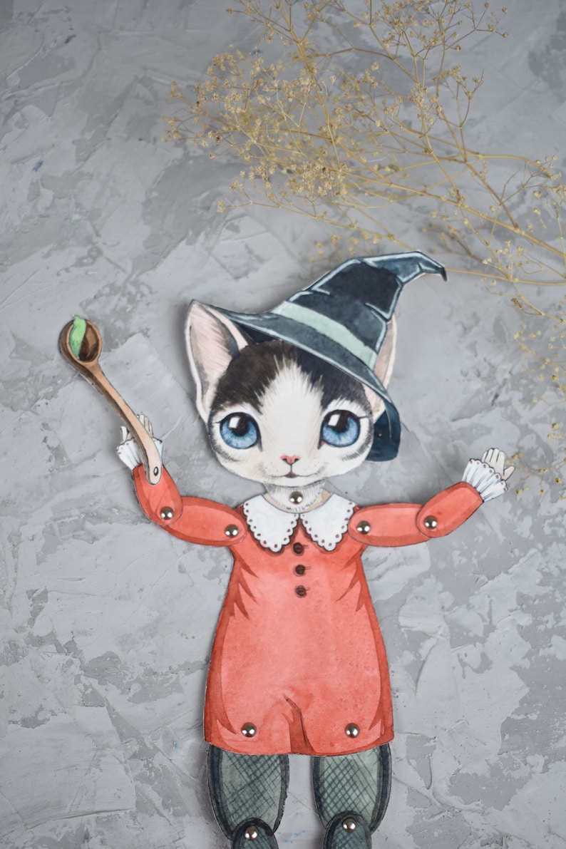 May include: A paper doll of a cat wearing a witch's hat and a red dress, holding a wooden spoon with a green ball on it. The doll is on a grey background with dried flowers.