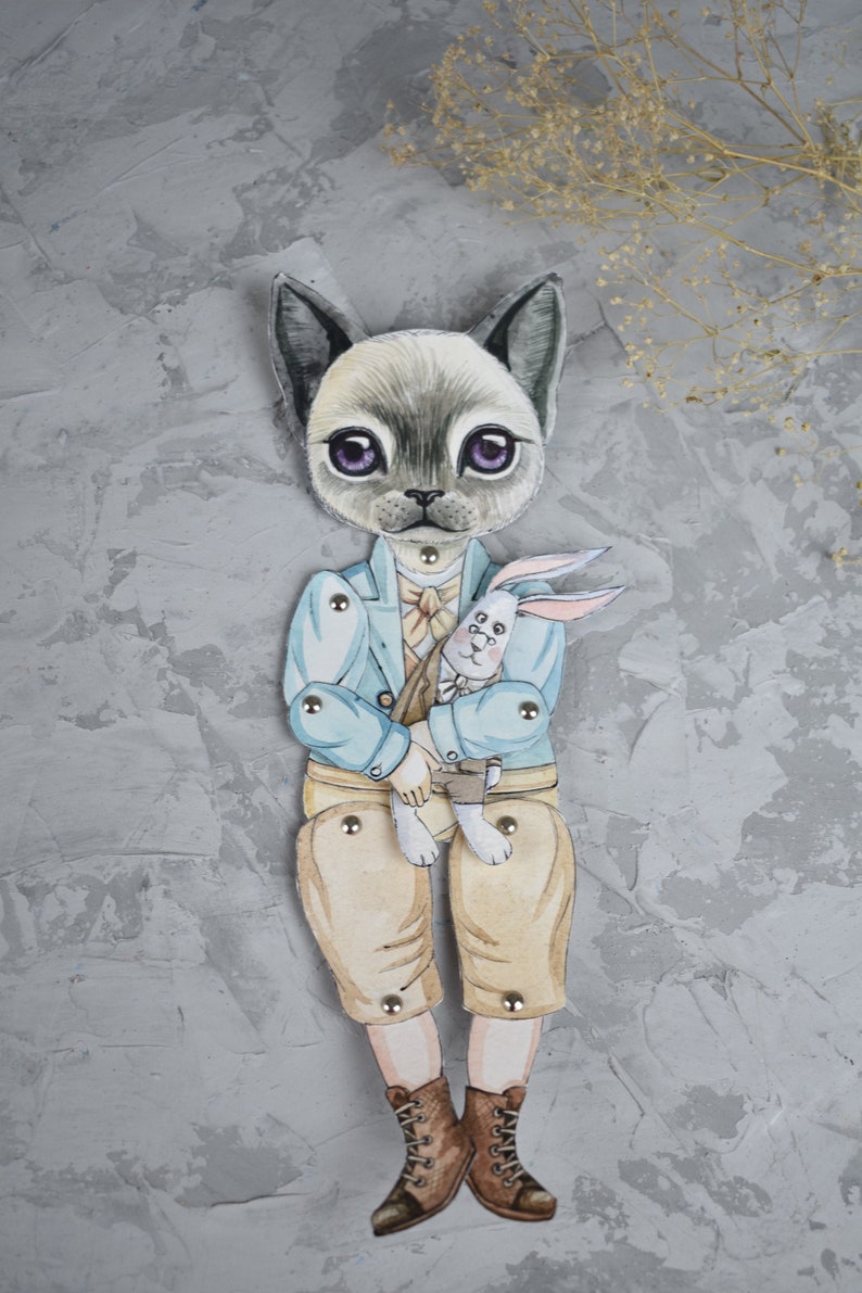 Puede incluir: Una mu&ntilde;eca de papel caprichosa con un personaje felino con una chaqueta azul claro, pantalones color canela y botas marrones, sosteniendo un conejo blanco. La mu&ntilde;eca tiene articulaciones y est&aacute; sobre un fondo gris texturizado.