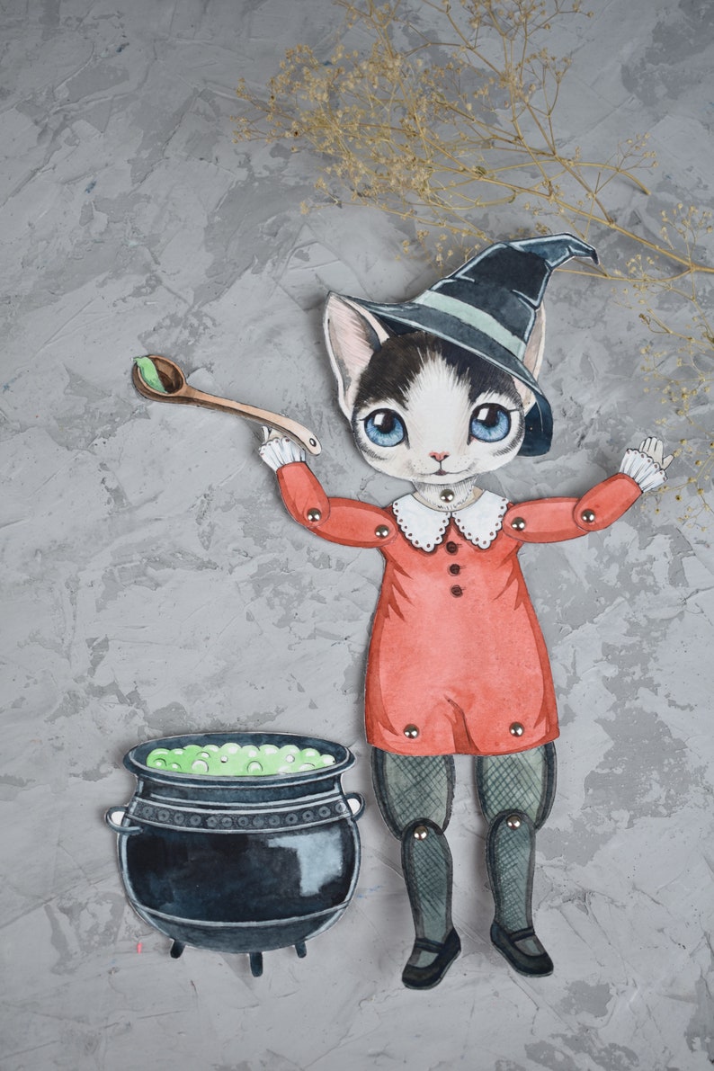 May include: A black and white cat wearing a witch's hat and red dress holds a wooden spoon over a cauldron filled with green liquid. The cat is a paper doll with movable arms and legs.