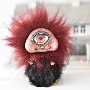 Op de afbeelding: Een klein, pluche, rood en zwart beestje met een groot oog en een glimlach. Het beestje is gemaakt van stof en heeft een geschilderd gezicht.