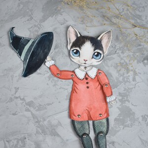 May include: A black and white cat dressed as a witch with a black pointed hat and a red dress. The cat is a paper doll with movable arms and legs.