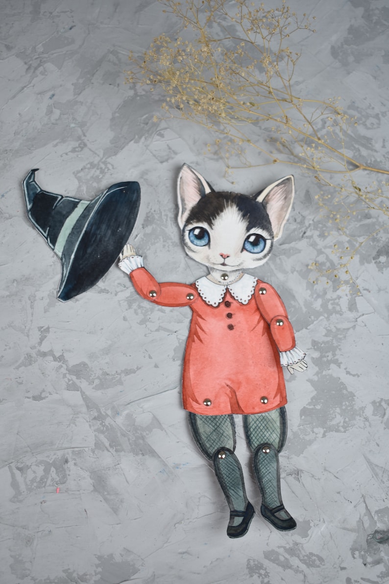 May include: A black and white cat dressed as a witch with a black pointed hat and a red dress. The cat is a paper doll with movable arms and legs.