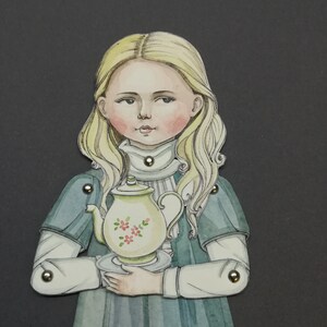 May include: A paper doll illustration of a girl with blonde hair, wearing a blue dress and holding a teapot. The teapot is cream-colored with pink floral accents. The doll has articulated joints with metal fasteners.