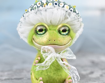 May include: A green frog plush toy wearing a white lace shower cap with green and gold polka dots. The frog has large green eyes and a pink smile.