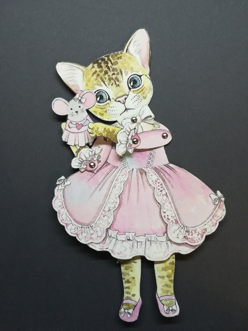 May include: A paper doll of a cat wearing a pink dress and holding a small mouse doll. The cat has brown and black spots and blue eyes. The dress has lace trim and a bow.