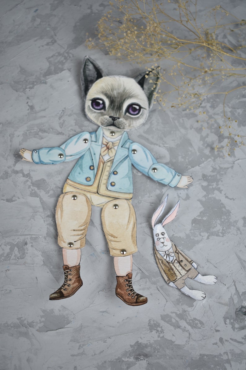 Puede incluir: Una mu&ntilde;eca de papel caprichosa con un personaje de gato con una chaqueta azul claro, pantalones color canela y botas marrones. Tambi&eacute;n hay una mu&ntilde;eca de conejo a juego. Las mu&ntilde;ecas est&aacute;n conectadas con sujetadores met&aacute;licos. Fondo gris texturizado.