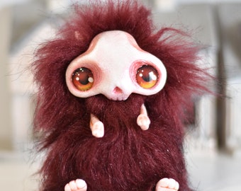 May include: A small, furry, red plush toy with a white skull-shaped head and large, orange eyes. The toy has a small, pink nose and a pink mouth.