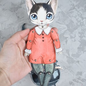 May include: A paper doll of a cat wearing a red dress with white trim and black tights. The doll is holding a black cauldron and has movable joints.