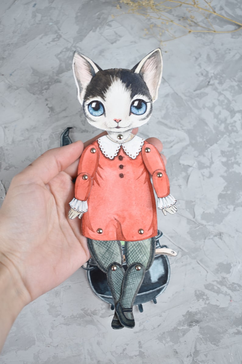 May include: A paper doll of a cat wearing a red dress with white trim and black tights. The doll is holding a black cauldron and has movable joints.