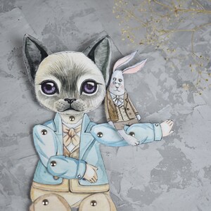 Puede incluir: Una mu&ntilde;eca de gato articulada y caprichosa, con una chaqueta azul claro y un conejo como compa&ntilde;ero. El gato tiene grandes ojos morados y sostiene al conejo. La mu&ntilde;eca est&aacute; hecha de papel y tiene remaches met&aacute;licos en las articulaciones.
