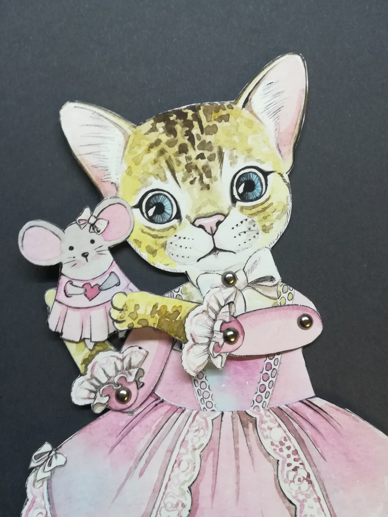 May include: A paper doll of a cat wearing a pink dress and holding a mouse doll. The cat has brown and white fur and blue eyes. The dress has lace trim and a floral pattern.