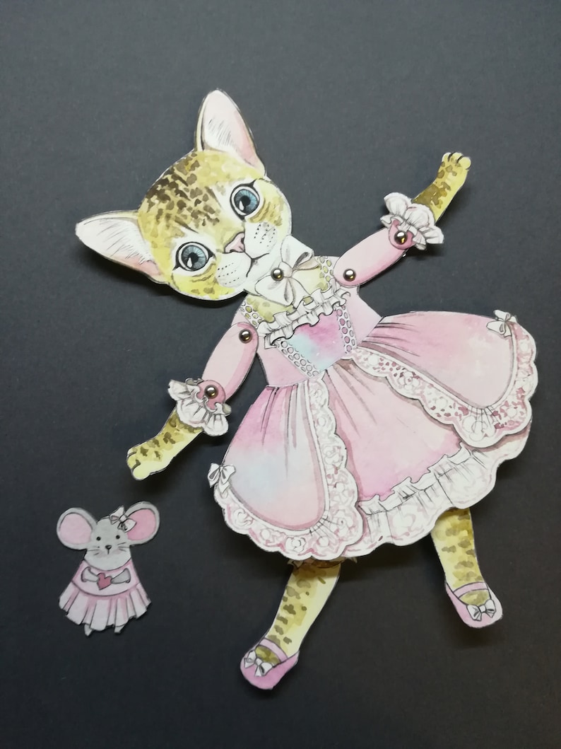 May include: A paper doll of a cat wearing a pink dress with lace trim and a white bow. The cat has brown and black spots and blue eyes. A smaller paper doll of a mouse wearing a pink dress with a heart is also included.