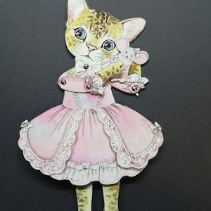 May include: A paper doll of a cat wearing a pink dress with lace trim and holding a small mouse doll. The cat has brown and white fur and blue eyes.