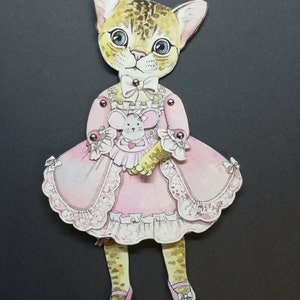 May include: A paper doll of a cat wearing a pink dress with lace trim and holding a small mouse doll. The cat has brown and black spots and blue eyes.